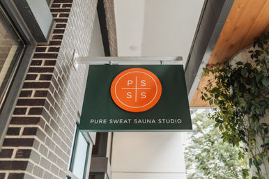 Green hanging storefront sign with a bright orange circular logo above a glass doorway on a brick facade, marking a sauna studio entrance