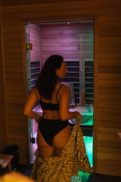 Person in black swimwear entering a wooden infrared sauna with purple and green mood lighting while holding a patterned towel
