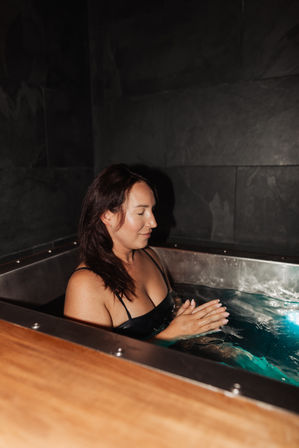 Woman relaxing in a stainless-steel cold plunge bath at an indoor spa, eyes closed and hands clasped amid dark tiled walls — hydrotherapy wellness scene.