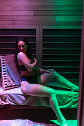 Person in black swimwear relaxing on a towel-covered wooden sauna bench under pink and green mood lighting in an indoor spa sauna.