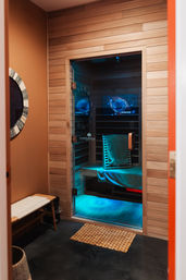 Cozy wood-paneled infrared sauna with glass door, blue LED glow, towel-draped bench and a small spa changing area with round mirror.