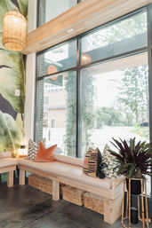 Cozy sunlit seating nook with a cushioned wooden bench, colorful patterned pillows, wicker storage baskets underneath, potted plant on a bamboo stand, and large floor-to-ceiling windows overlooking a tree-lined urban street.