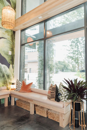 Cozy sunlit seating nook with a cushioned wooden bench, colorful patterned pillows, wicker storage baskets underneath, potted plant on a bamboo stand, and large floor-to-ceiling windows overlooking a tree-lined urban street.