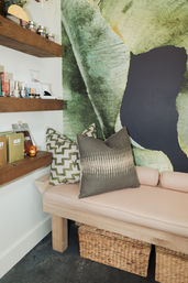 Cozy spa-style seating nook with pale pink cushioned wooden bench, patterned green and gray throw pillows, large green botanical mural, wooden shelves displaying skincare products and a candle, and woven storage baskets underneath.