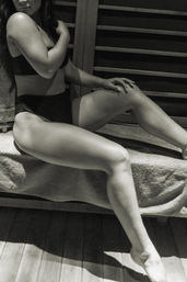 Black-and-white photo of a person in black swimwear reclining on a towel-covered wooden sauna bench, legs extended and hand resting on knee, spa relaxation vibe