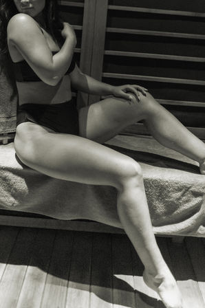 Black-and-white photo of a person in black swimwear reclining on a towel-covered wooden sauna bench, legs extended and hand resting on knee, spa relaxation vibe