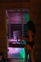 Woman in black swimwear opening a glass door to a wooden infrared sauna with colorful LED mood lighting and a towel-draped bench.