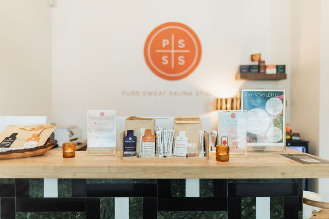 Bright sauna studio reception desk with wellness product display, amber candles, a newsletter sign, and a round orange logo on a minimalist white wall.