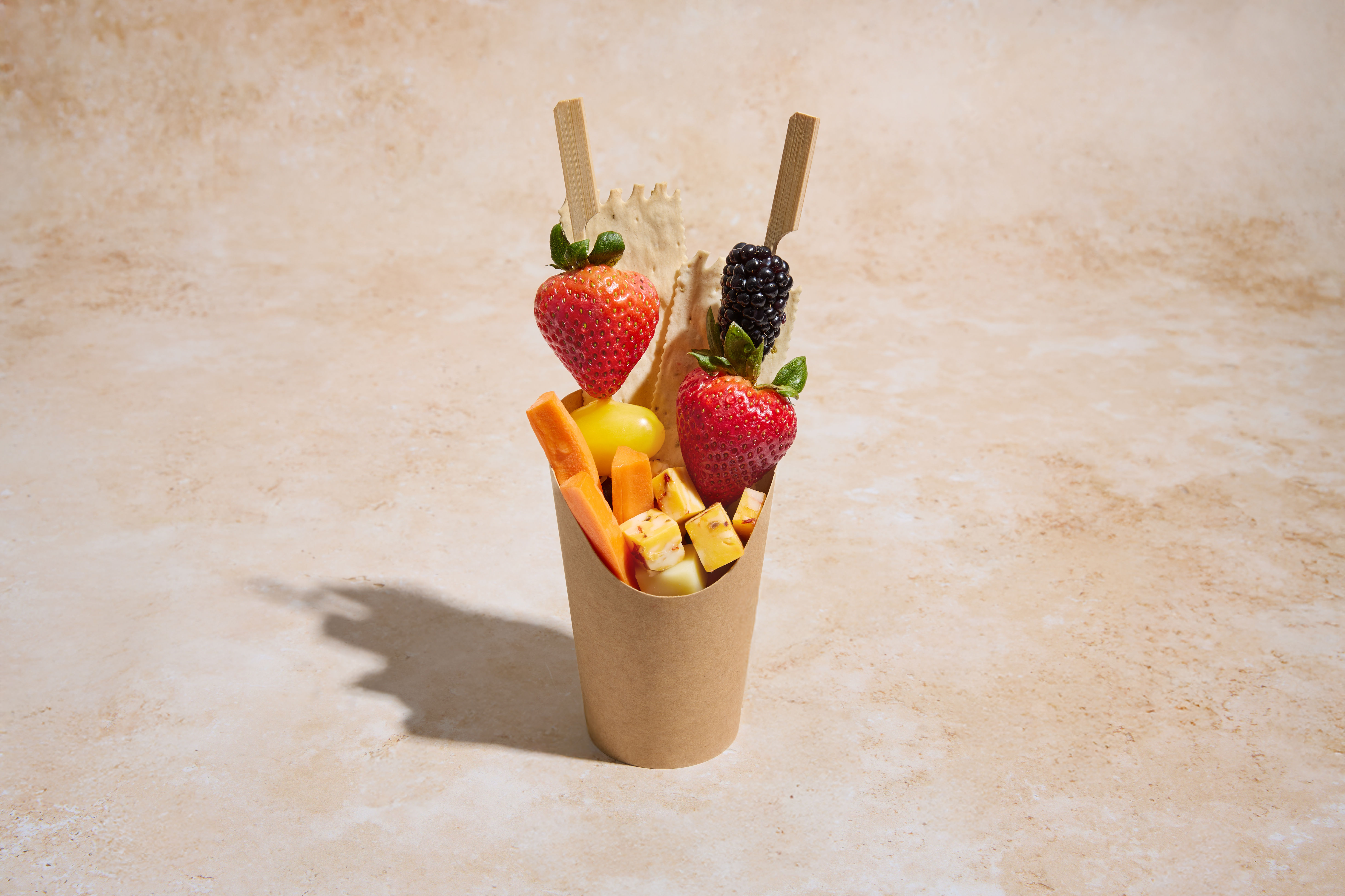 Brown paper cup holding a fresh snack: two wooden skewers with strawberries and a blackberry, grilled pineapple cubes, carrot sticks, a yellow cherry tomato and a crisp cracker on a neutral beige surface.