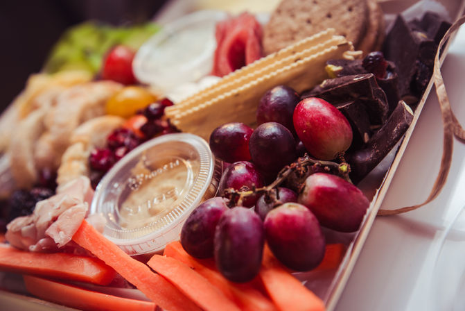 Close-up of a vibrant charcuterie box with red grapes, carrot sticks, assorted crackers, chocolate chunks, cured meats and dipping sauces