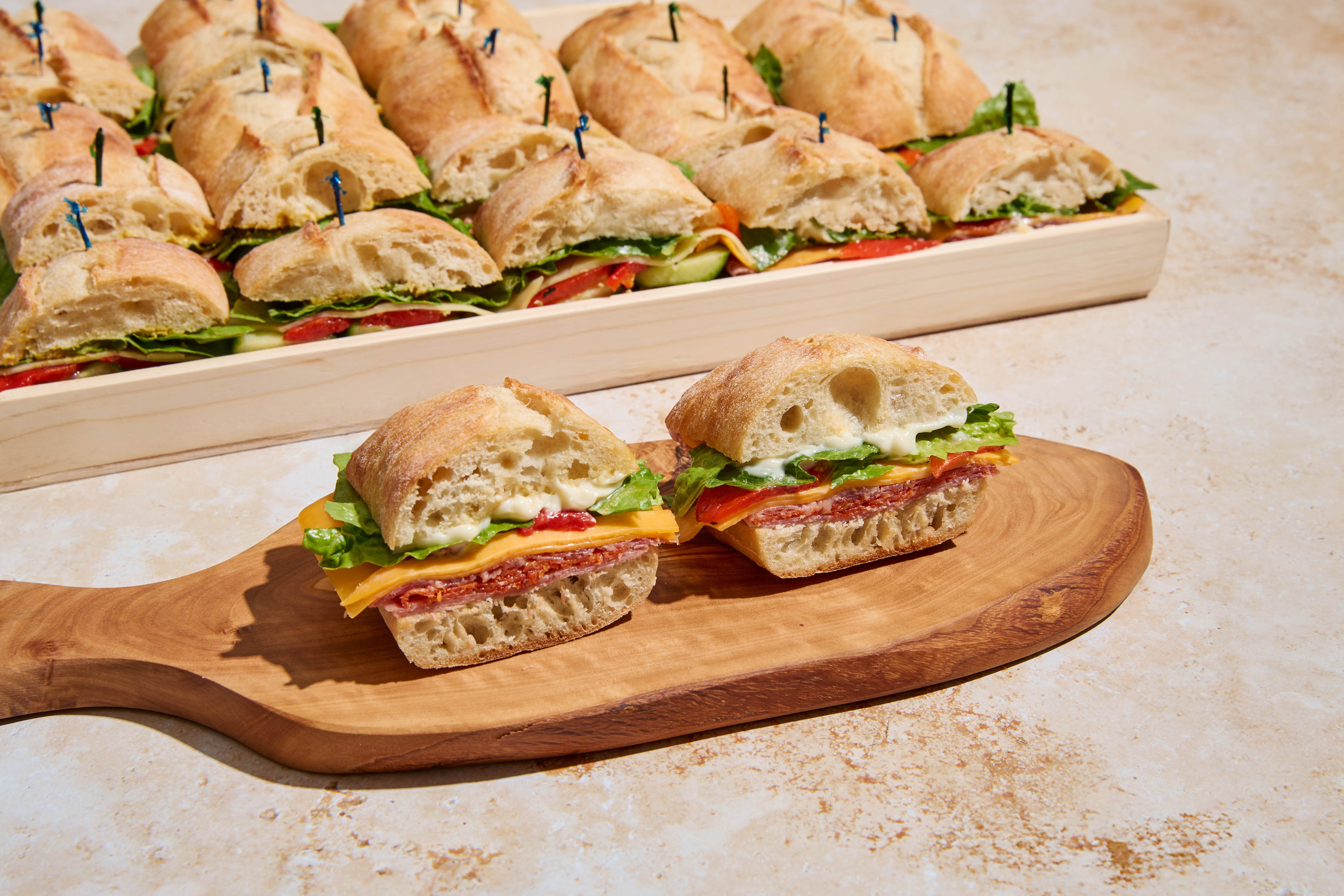 Bite-size ciabatta sandwiches with salami, cheddar, lettuce and mayo on a wooden board, with a full catering platter of sliders in the background