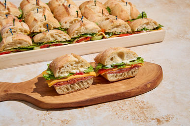 Bite-size ciabatta sandwiches with salami, cheddar, lettuce and mayo on a wooden board, with a full catering platter of sliders in the background