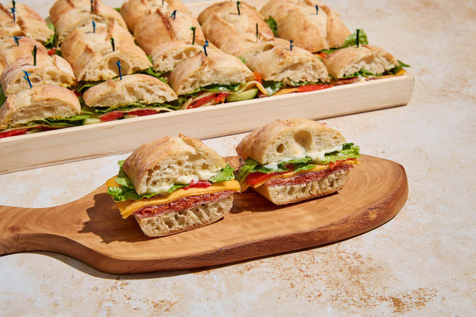 Bite-size ciabatta sandwiches with salami, cheddar, lettuce and mayo on a wooden board, with a full catering platter of sliders in the background