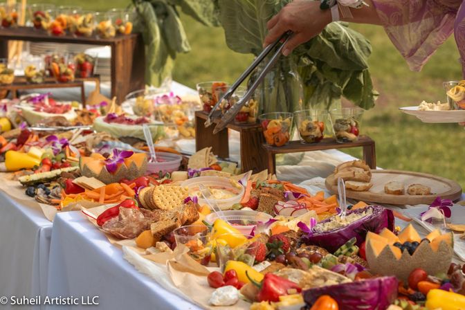 Outdoor garden buffet with colorful grazing-table spread of charcuterie, cheeses, crackers, fresh fruit, vegetables, dips and edible flowers, a hand using tongs to serve onto a plate.