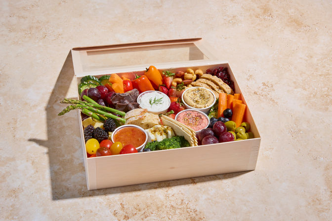 Wooden gourmet grazing box with colorful fresh fruit (grapes, blackberries, cherry tomatoes), raw vegetables (carrots, asparagus, broccoli), assorted crackers and cheeses, olives, nuts, and three creamy dips, styled for picnic or appetizer spread.