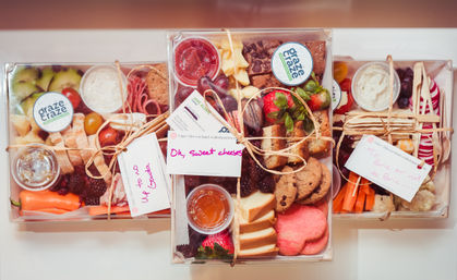 Three tied clear grazing boxes of charcuterie-style snacks — strawberries, grapes, apple slices, cookies, brownies, crackers, sliced bread, cheeses, sliced salami, carrot and cauliflower crudités, small dip cups and handwritten gift tags on a white surface.