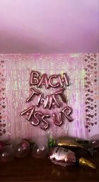 Pink iridescent party backdrop with rose-gold foil letter balloons reading BACK THAT ASS UP, heart-patterned side walls, and scattered clear and gold balloons on the floor.