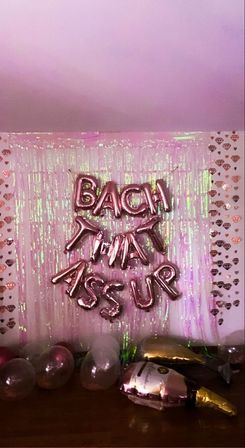 Pink iridescent party backdrop with rose-gold foil letter balloons reading BACK THAT ASS UP, heart-patterned side walls, and scattered clear and gold balloons on the floor.