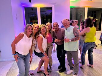 Smiling group of six adults posing with drinks at an indoor party under colorful LED lighting, casual summer outfits and wood‑look floors