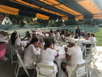 Group of tennis players in white outfits enjoying lunch at round tables on a shaded clubhouse terrace with an orange awning, overlooking outdoor tennis courts.
