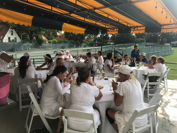Group of tennis players in white outfits enjoying lunch at round tables on a shaded clubhouse terrace with an orange awning, overlooking outdoor tennis courts.
