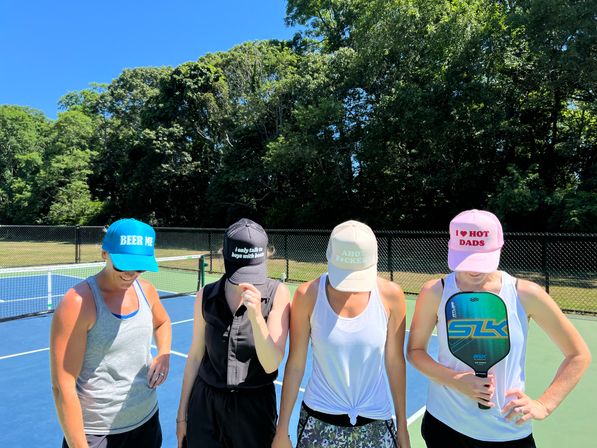 Four players on a sunny outdoor pickleball court wearing colorful slogan caps and athletic gear, one holding a pickleball paddle, fenced court and tree-lined park in the background.