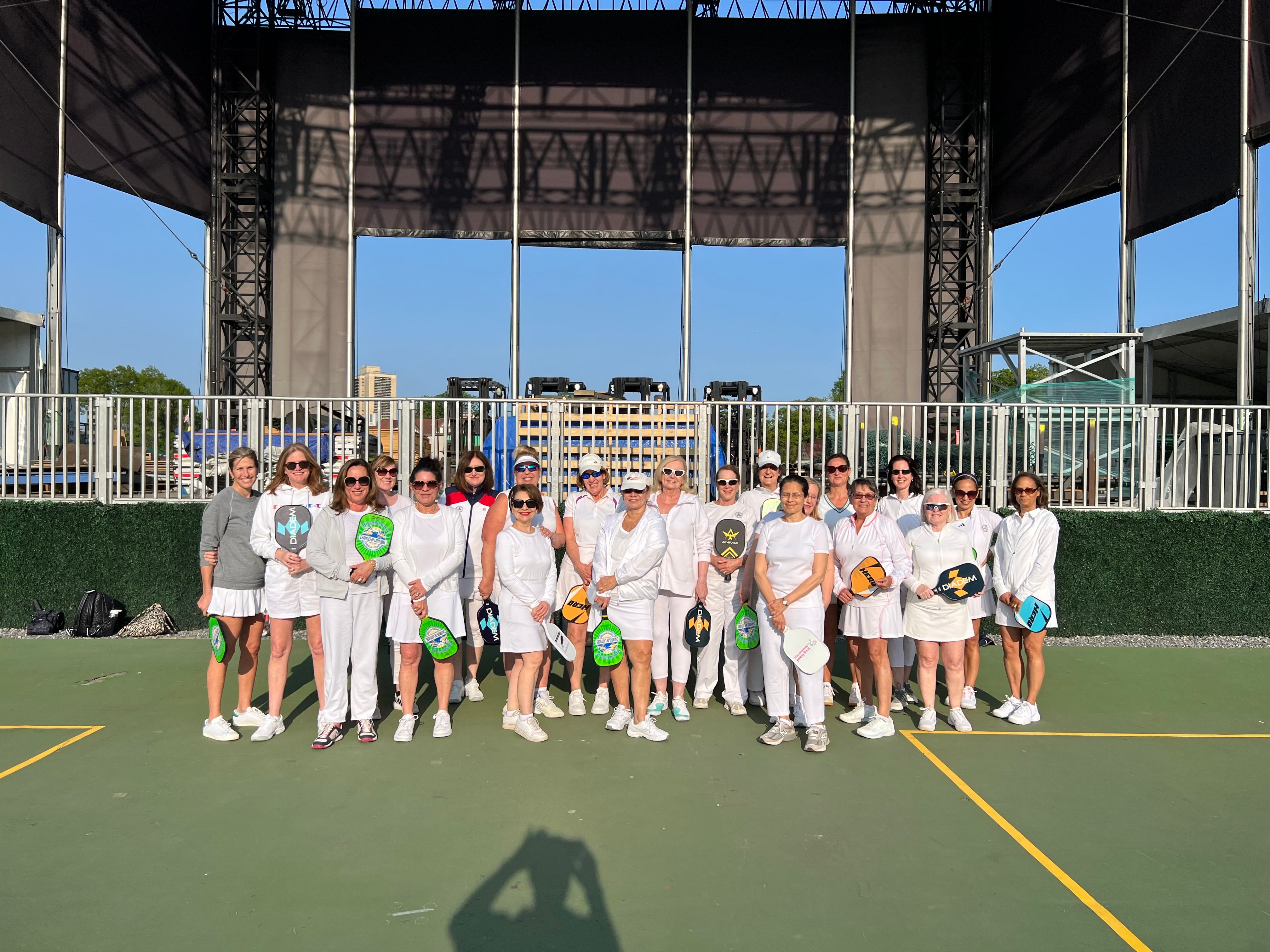 Group of women in white athletic outfits posing with colorful pickleball paddles on a sunny outdoor court in front of a large stage