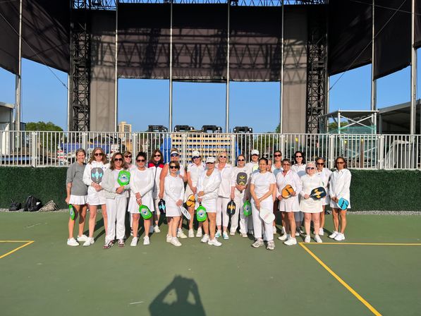 Group of women in white athletic outfits posing with colorful pickleball paddles on a sunny outdoor court in front of a large stage