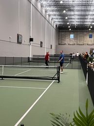 Indoor pickleball courts at a recreational sports center with green courts, nets, high industrial ceiling and players warming up