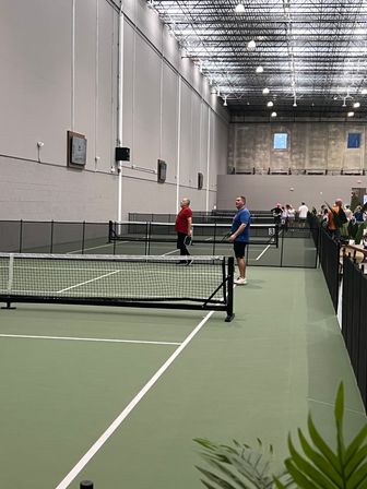 Indoor pickleball courts at a recreational sports center with green courts, nets, high industrial ceiling and players warming up