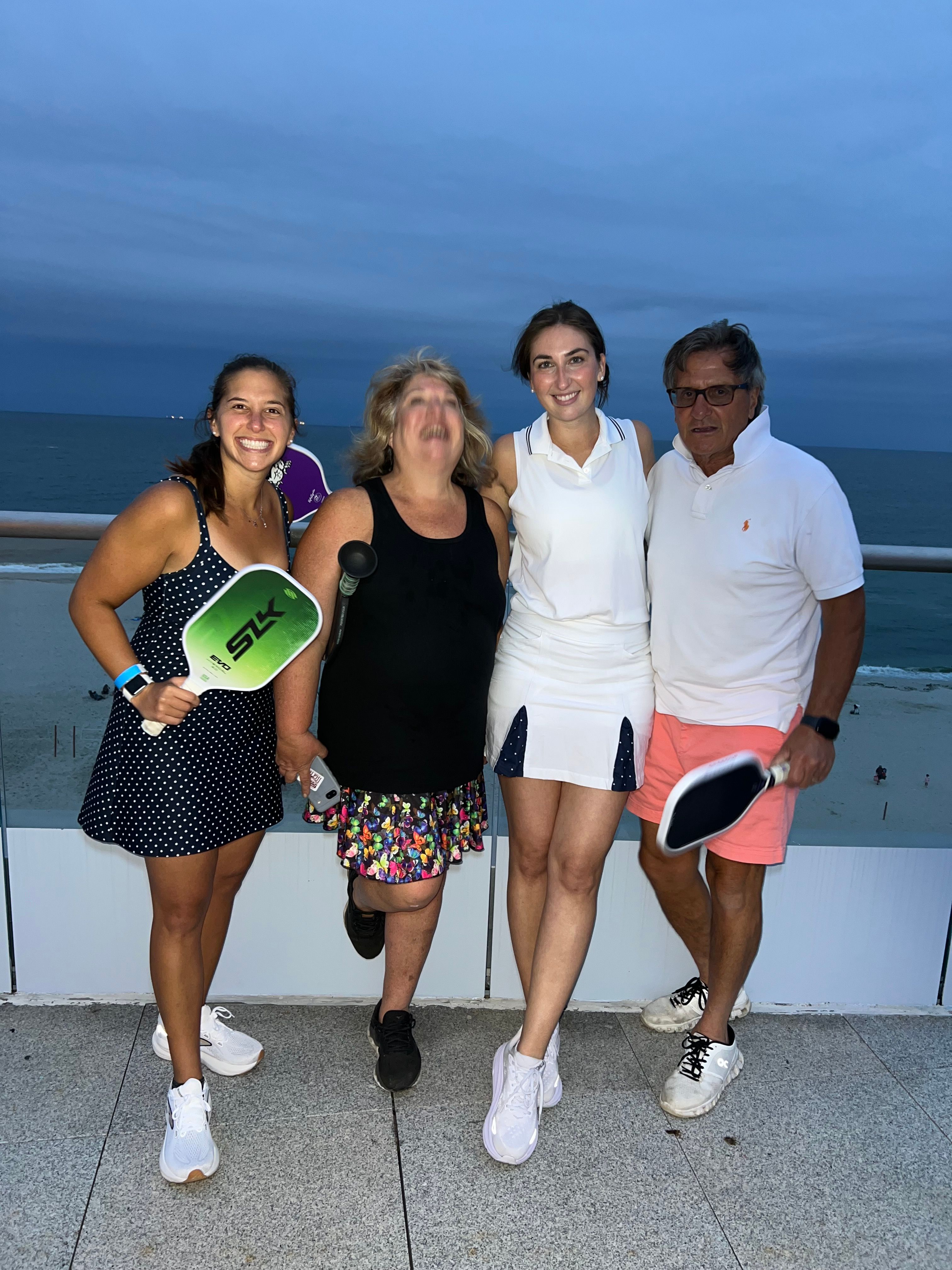 Four adults on an oceanfront balcony at twilight, smiling and posing with pickleball paddles and sneakers, sandy beach and calm sea in the background.
