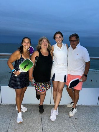 Four adults posing on an oceanfront balcony at dusk, smiling in summer outfits and holding pickleball paddles with the beach and calm sea in the background.