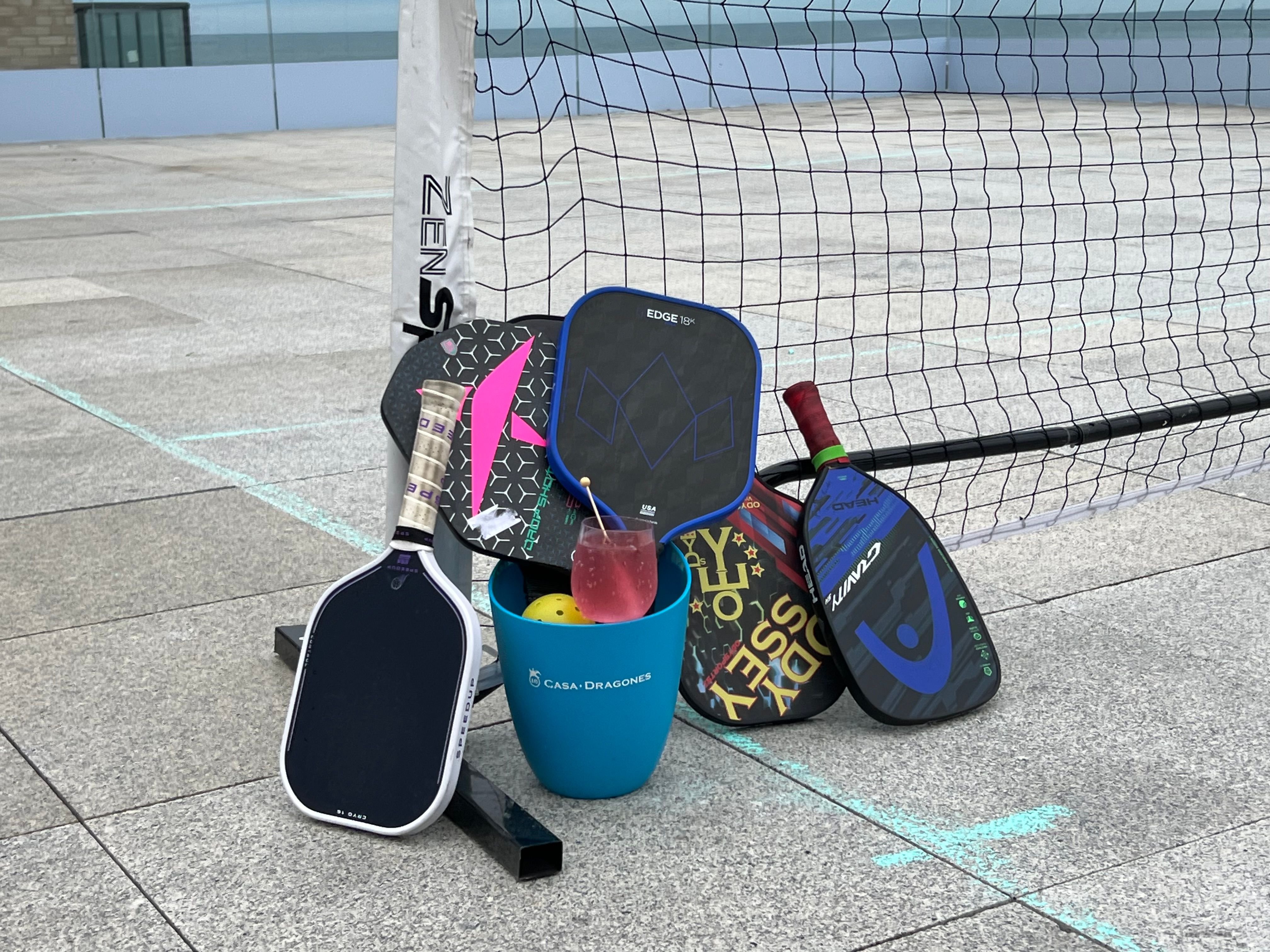 Colorful pickleball paddles leaning against a net post on an outdoor concrete court, next to a blue bucket holding a ball and a pink drink.