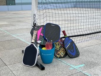 Colorful pickleball paddles leaning against a net post on an outdoor concrete court, next to a blue bucket holding a ball and a pink drink.