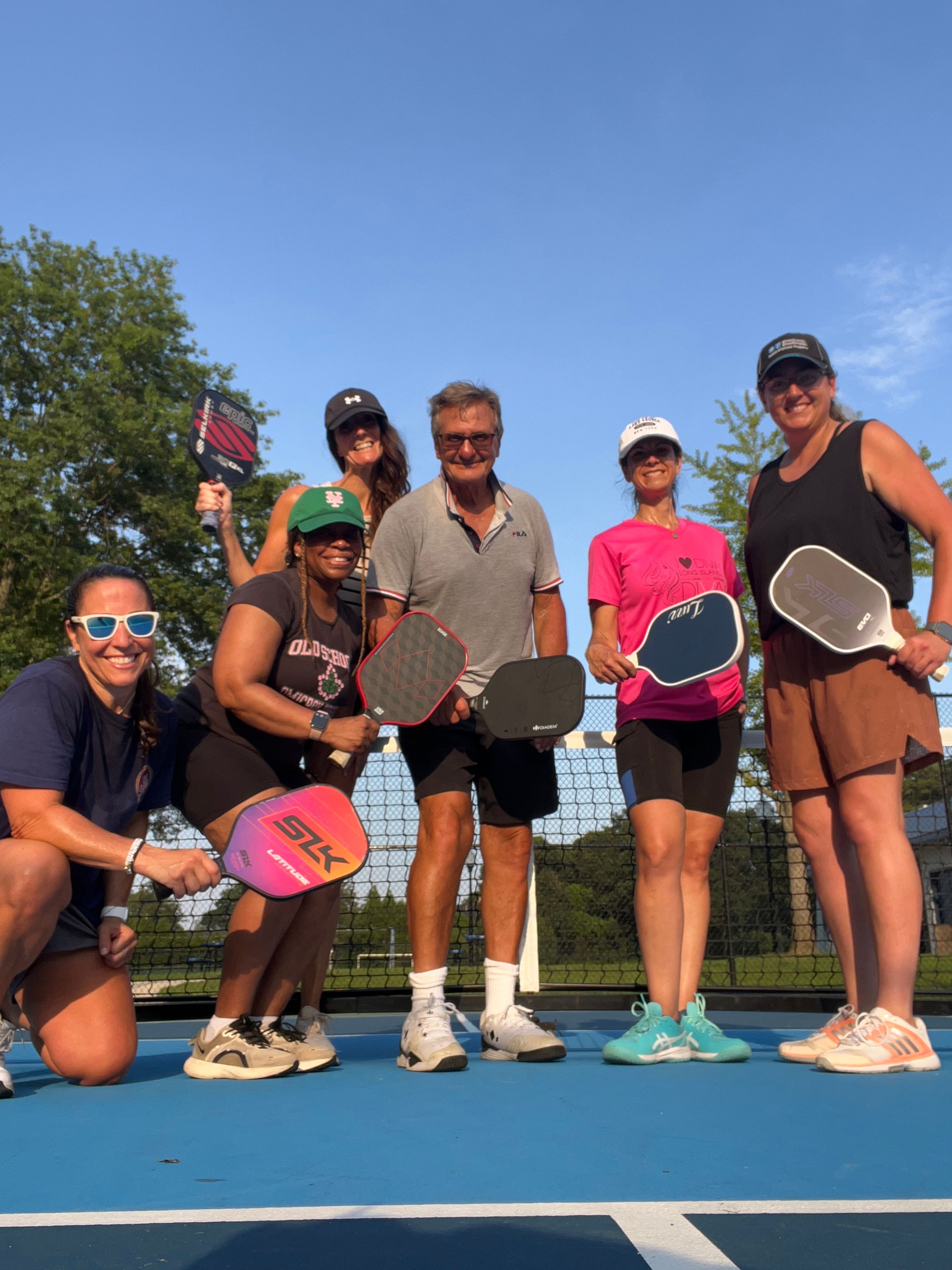 Six adults smiling on a blue outdoor pickleball court, posing with paddles under a clear blue sky in athletic gear in front of the net and trees
