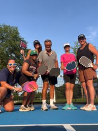 Six adults smiling on a blue outdoor pickleball court, posing with paddles under a clear blue sky in athletic gear in front of the net and trees