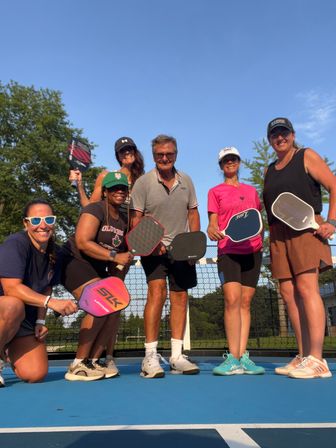 Six adults smiling on a blue outdoor pickleball court, posing with paddles under a clear blue sky in athletic gear in front of the net and trees