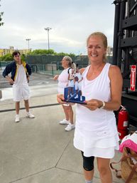 Smiling woman in white tennis outfit holding a small plaque of two players, standing by outdoor tennis courts with teammates and city buildings in the background