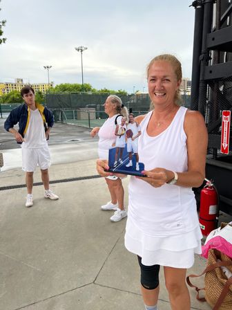 Smiling woman in white tennis outfit holding a small plaque of two players, standing by outdoor tennis courts with teammates and city buildings in the background