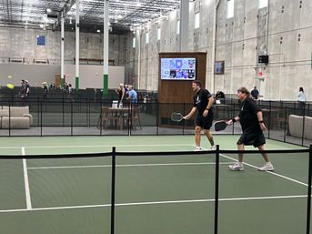 Two players ready at the net on an indoor pickleball court as a yellow ball arcs by, with multiple courts, spectators, and a lounge area visible behind a black mesh divider.