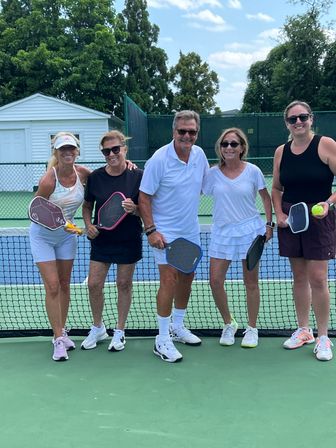 Five smiling pickleball players at the net on an outdoor green court, holding paddles and a yellow ball with trees and blue sky in the background.