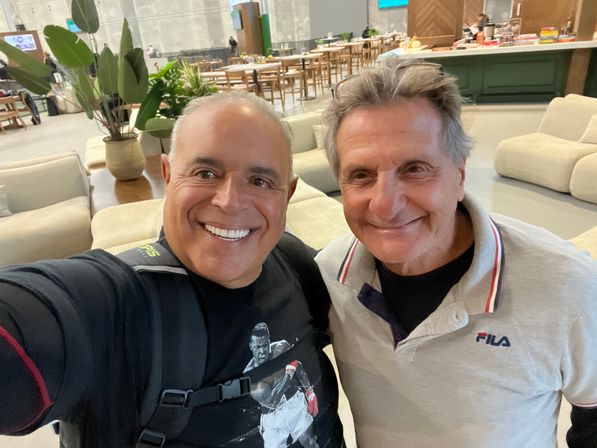 Two smiling men take a selfie in a bright modern lounge with comfy sofas, potted plants and communal wooden tables — casual indoor meet-up scene.
