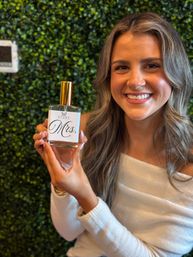 Smiling woman holding a perfume bottle labeled “Mrs.” against a green boxwood backdrop, wearing a white off-shoulder top — bridal fragrance product shot.