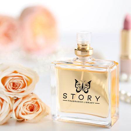 Square glass perfume bottle with golden liquid and butterfly logo on a white vanity, surrounded by peach roses and a pink lipstick