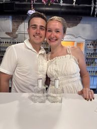 Smiling young couple in white outfits posing behind a counter with two clear perfume bottles in a bright fragrance boutique with shelves of bottles and exposed brick wall