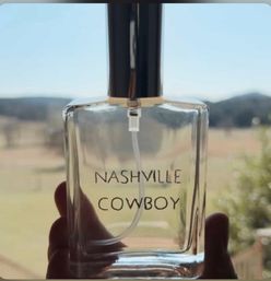 Clear glass perfume spray bottle labeled 'NASHVILLE COWBOY' held up to a sunlit, blurred countryside with distant hills