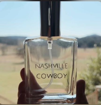 Clear glass perfume spray bottle labeled 'NASHVILLE COWBOY' held up to a sunlit, blurred countryside with distant hills
