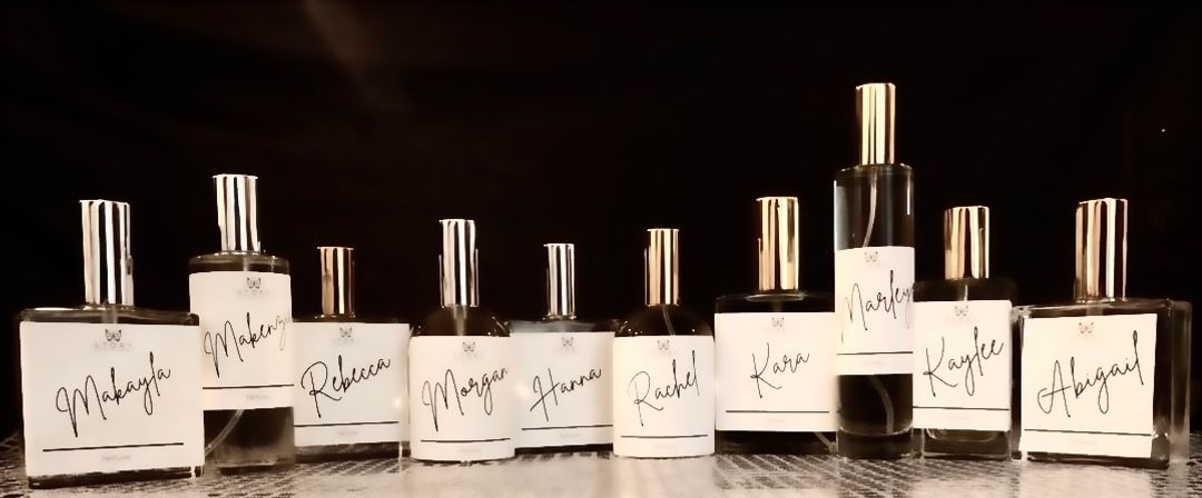 Chic display of glass perfume bottles with white handwritten name labels and gold caps arranged on a reflective surface against a black backdrop — boutique-style fragrance display