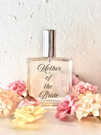 Perfume bottle labeled Mother of the Bride on a white textured backdrop, surrounded by pastel pink and cream flowers — elegant wedding gift or bridal-suite accessory.