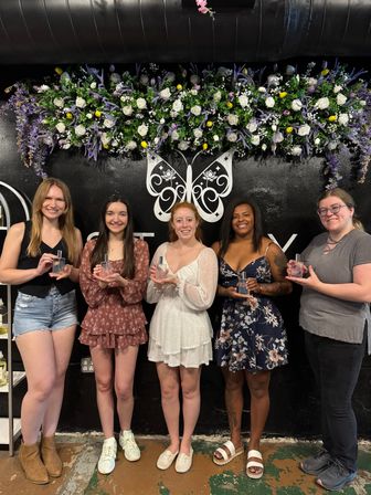 Five smiling women holding glass perfume bottles in a boutique fragrance workshop, posed in front of a black wall with a white butterfly logo and purple-and-white floral garland backdrop.